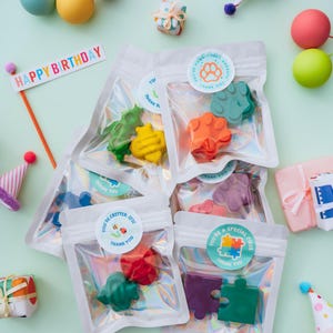 May include: Several clear, iridescent pouches containing shaped crayons in various colours. Each pouch features a round sticker with text such as 'You're Pawsitively Awesome' or 'You're Critter-ific'. A 'Happy Birthday' sign and small party hats are visible.