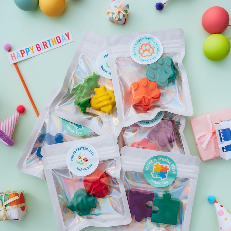 May include: Several clear, iridescent pouches containing shaped crayons in various colours. Each pouch features a round sticker with text such as 'You're Pawsitively Awesome' or 'You're Critter-ific'. A 'Happy Birthday' sign and small party hats are visible.