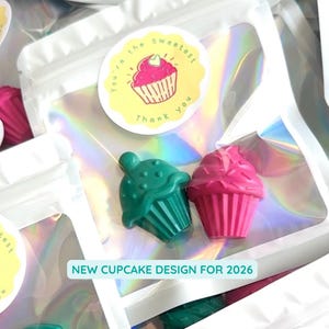 May include: A holographic pouch containing two cupcake-shaped wax crayons, one dark green and one bright pink. A circular sticker on the front features a pink cupcake illustration with the text "You're the sweetest Thank you" and "NEW CUPCUPCAKE DESIGN FOR 2026".