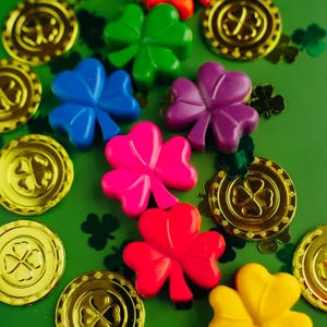 May include: A collection of colorful plastic shamrock shapes in green, blue, purple, pink, red, and yellow, interspersed with metallic gold coins featuring a four-leaf clover design on a solid green background, creating a festive decorative arrangement.