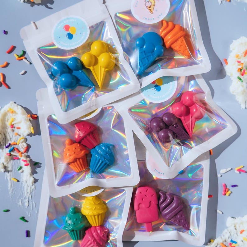 May include: Five iridescent pouches containing shaped wax crayons. Each pouch holds two distinct figures, including ice cream cones, cupcakes, and popsicles in vibrant colours like blue, yellow, orange, pink, purple, and green, with circular stickers.