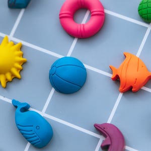 May include: A collection of small, colourful silicone shapes including a bright pink life preserver, a yellow sun, a blue beach ball, an orange fish, a blue whale, a purple dolphin, and a green textured piece, all arranged on a light blue grid.