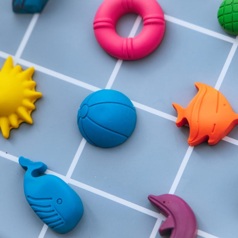 May include: A collection of small, colourful silicone shapes including a bright pink life preserver, a yellow sun, a blue beach ball, an orange fish, a blue whale, a purple dolphin, and a green textured piece, all arranged on a light blue grid.