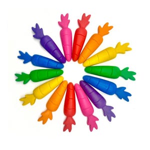 May include: A collection of fourteen carrot-shaped crayons arranged in a circular pattern. Each crayon features a distinct solid color including red, orange, yellow, green, blue, purple, and pink, with textured horizontal lines along the body.