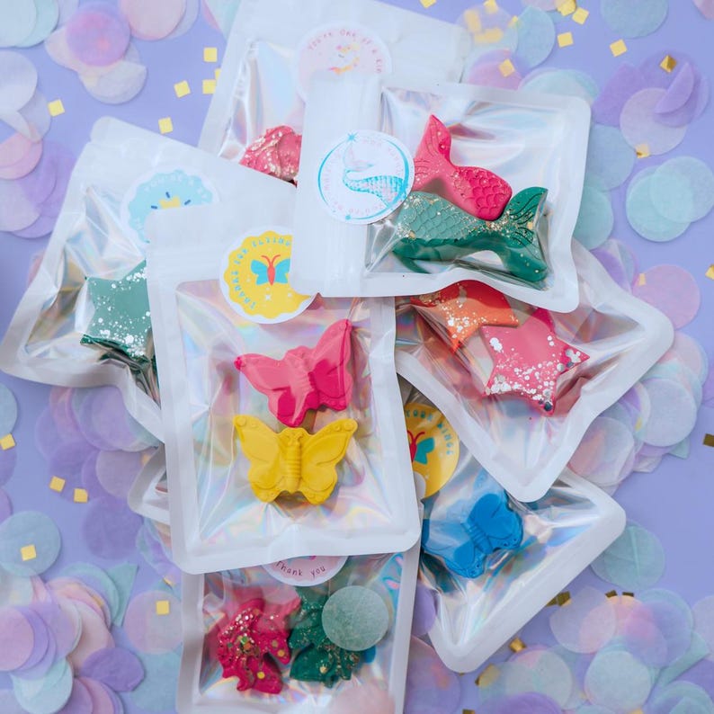 May include: A collection of small, shaped wax crayons packaged in individual clear, resealable pouches. The crayons feature various designs including butterflies, stars, and mermaid tails in vibrant colours like pink, yellow, green, and blue.