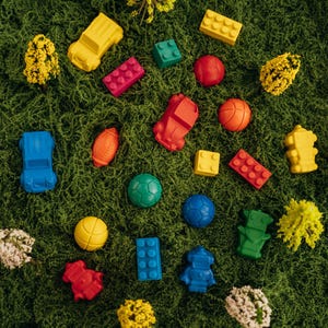 May include: A collection of colourful plastic toy shapes including cars, building blocks, robots, and various sports balls scattered across a textured green surface, interspersed with small yellow and white decorative floral accents.