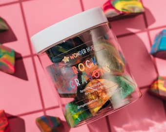 May include: A clear plastic jar with a white lid containing multi-colored rock-shaped crayons. The label reads WONDER HUE ROCK CRAYONS, Sparking imagination thru color + play, and @mywonderhue.com. The crayons feature swirls of blue, green, red, and yellow.