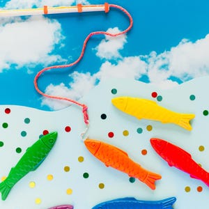 Könnte beinhalten: Ein farbenfrohes Angelspiel vor einem blauen Himmel mit weißen Wolken. Das Set enthält eine Angelrute und Spielzeugfische in verschiedenen Farben: Gelb, Rot, Orange, Grün, Blau und Lila. Die Szene ist mit bunten Konfetti dekoriert.