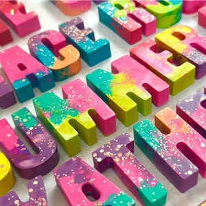 May include: Colorful, block-style letters spelling "SUMMER" in various vibrant hues. Each letter is a different color combination, including pink, blue, yellow, and purple, with glitter accents. The letters are arranged in rows.
