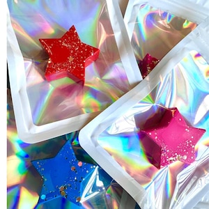 以下が含まれることがあります： 赤、ピンク、青の星型ワックスアイテム3点。それぞれに金色のグリッターが埋め込まれています。各アイテムは、虹色のスペクトルを反射する透明なホログラフィック加工のポーチに個別に密封されています。