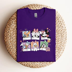 May include: A purple crew neck t-shirt featuring a graphic print of six polaroid-style frames. Each frame contains a different Disney character: Pluto, Minnie Mouse, Mickey Mouse, Donald Duck, Daisy Duck, and Goofy, with their names written below.