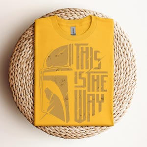 May include: Mustard yellow t-shirt with a distressed graphic of a helmet and the text "THIS IS THE WAY" in a vertical layout. The shirt is folded and placed on a round woven mat.