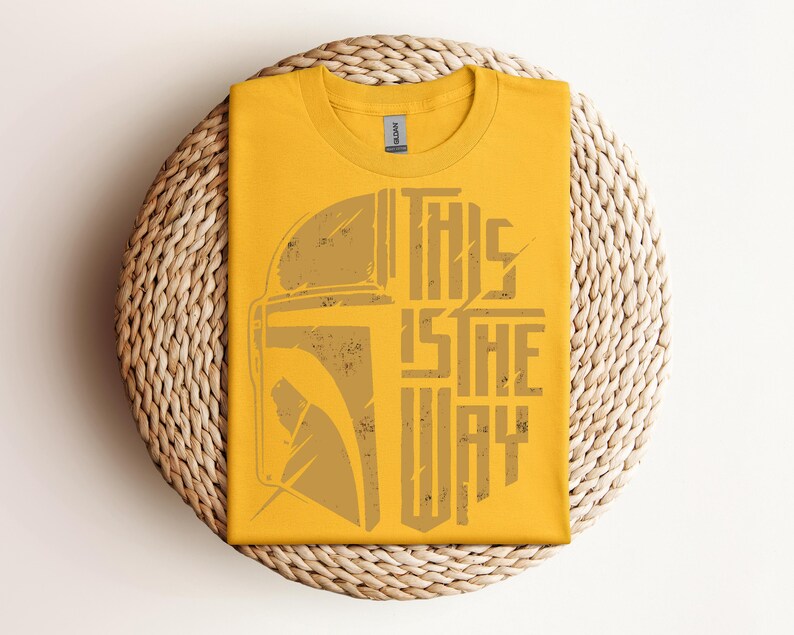 May include: Mustard yellow t-shirt with a distressed graphic of a helmet and the text "THIS IS THE WAY" in a vertical layout. The shirt is folded and placed on a round woven mat.