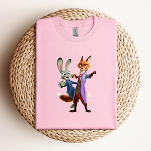 May include: A light pink t-shirt featuring a cartoon image of Judy Hopps and Nick Wilde from Zootopia. Judy is a gray rabbit wearing a blue jacket, and Nick is an orange fox in a purple coat. The shirt is folded on a woven mat.
