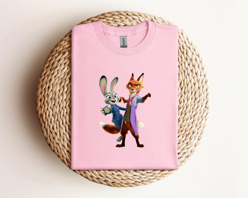 May include: A light pink t-shirt featuring a cartoon image of Judy Hopps and Nick Wilde from Zootopia. Judy is a gray rabbit wearing a blue jacket, and Nick is an orange fox in a purple coat. The shirt is folded on a woven mat.