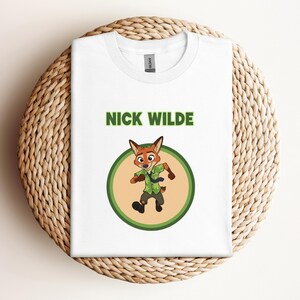 May include: White t-shirt featuring a cartoon image of Nick Wilde from Zootopia. The design includes the character's image inside a green circle, with the name "NICK WILDE" printed above in green letters. The shirt is folded on a woven, light brown surface.