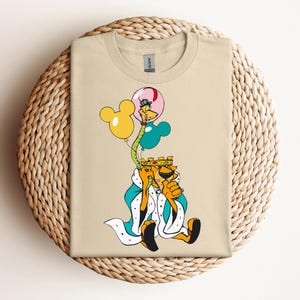 Pode incluir: Uma camiseta bege com uma ilustração de desenho animado de um leão usando uma coroa e segurando balões. Os balões são em forma de orelhas do Mickey Mouse e são amarelos, azuis e rosa.