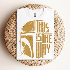 May include: White t-shirt with a gold graphic of a helmet and the text "THIS IS THE WAY". The shirt is folded and placed on a woven, circular surface. The design has a distressed, vintage look.