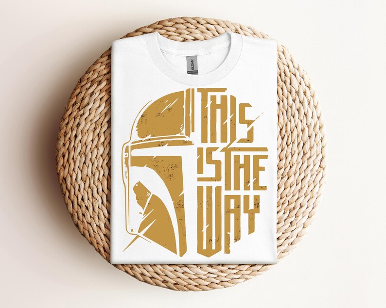 May include: White t-shirt with a gold graphic of a helmet and the text "THIS IS THE WAY". The shirt is folded and placed on a woven, circular surface. The design has a distressed, vintage look.