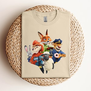 May include: A beige t-shirt featuring a colorful graphic of animated animal characters. The design includes a fox, a rabbit, a sheep, and a cheetah, all in vibrant colors. The shirt is folded on a woven, circular surface.