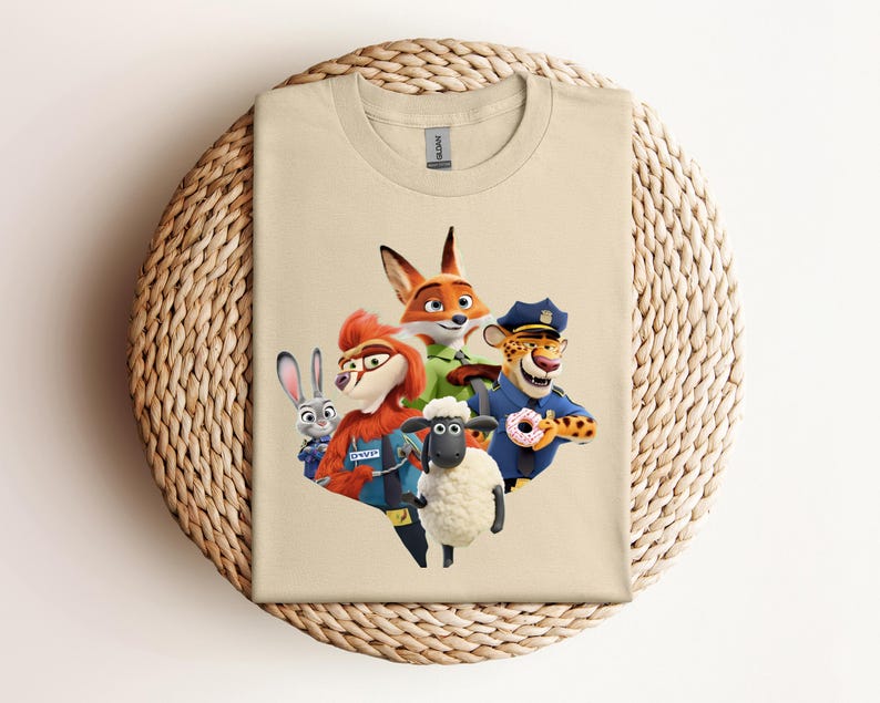 May include: A beige t-shirt featuring a colorful graphic of animated animal characters. The design includes a fox, a rabbit, a sheep, and a cheetah, all in vibrant colors. The shirt is folded on a woven, circular surface.