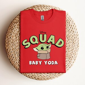 May include: A red crew neck t-shirt featuring the text "Squad" in green letters with a black outline, and the words "Baby Yoda" in white. The shirt has a graphic of Baby Yoda from the Star Wars series.