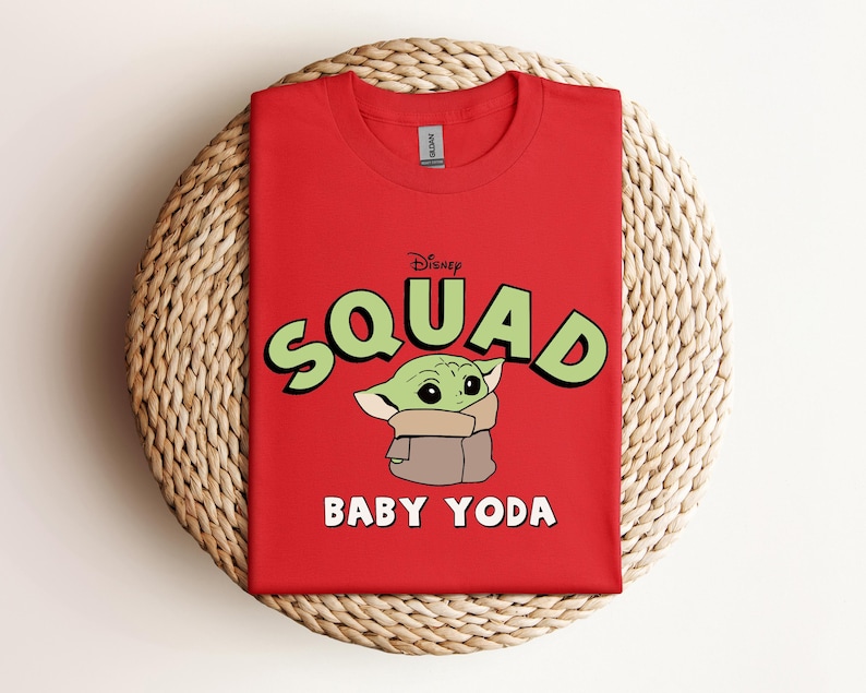 May include: A red crew neck t-shirt featuring the text "Squad" in green letters with a black outline, and the words "Baby Yoda" in white. The shirt has a graphic of Baby Yoda from the Star Wars series.