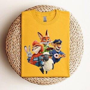 May include: Mustard yellow t-shirt featuring a colorful graphic of characters from the animated movie Zootopia. The design includes a fox, a rabbit, a sheep, a cheetah, and a character wearing a police hat. The shirt is displayed on a woven surface.