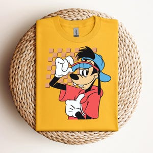 May include: A yellow t-shirt with a cartoon image of Goofy wearing a blue baseball cap, sunglasses, and a red shirt. The background is a checkered pattern.