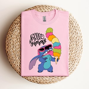 May include: A light pink t-shirt featuring a cartoon character wearing sunglasses and holding a colorful ice cream cone. The text "hello summer" is written above the character in black script.