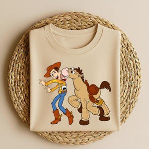 Peut inclure: T-shirt beige &agrave; col rond avec une illustration de dessin anim&eacute; d'un cow-boy et d'un cheval. Le cow-boy porte un chapeau, une chemise jaune, un pantalon bleu et des bottes marron. Le cheval a une selle et embrasse le cow-boy.