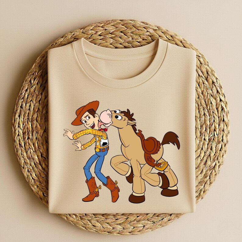 Peut inclure: T-shirt beige &agrave; col rond avec une illustration de dessin anim&eacute; d'un cow-boy et d'un cheval. Le cow-boy porte un chapeau, une chemise jaune, un pantalon bleu et des bottes marron. Le cheval a une selle et embrasse le cow-boy.