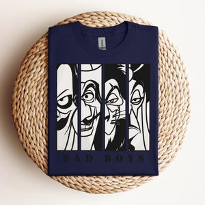 May include: Navy blue t-shirt featuring a graphic of four stylized villain faces in black and white vertical panels. The text "BAD BOYS" is printed in black below the faces. The shirt is folded on a woven surface.