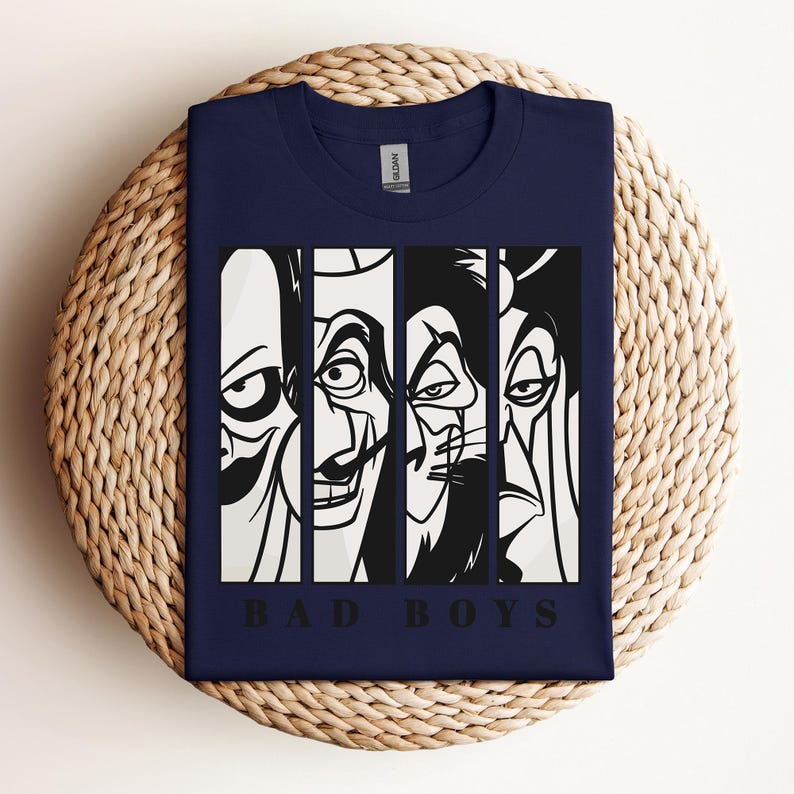 May include: Navy blue t-shirt featuring a graphic of four stylized villain faces in black and white vertical panels. The text "BAD BOYS" is printed in black below the faces. The shirt is folded on a woven surface.