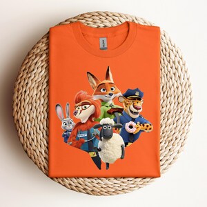 May include: An orange t-shirt featuring a colorful graphic of characters from the animated movie Zootopia. The design includes a fox, a rabbit, a sheep, a cheetah, and a chicken. The shirt is displayed on a woven, circular surface.