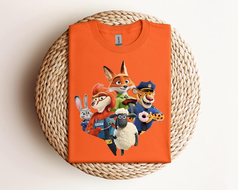 May include: An orange t-shirt featuring a colorful graphic of characters from the animated movie Zootopia. The design includes a fox, a rabbit, a sheep, a cheetah, and a chicken. The shirt is displayed on a woven, circular surface.