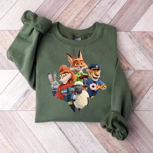 Op de afbeelding: Een groene sweatshirt met een kleurrijke afbeelding van personages uit de animatiefilm Zootopia. De afbeelding bevat een vos, een konijn, een schaap, een cheetah en andere personages. De sweatshirt heeft lange mouwen en een ronde hals.