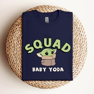 May include: Navy blue t-shirt featuring a graphic of Baby Yoda from the Disney+ series, The Mandalorian. The word "SQUAD" is in green letters with a black outline, and "BABY YODA" is in white. The shirt is displayed on a woven, circular surface.