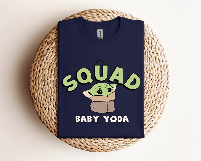 May include: Navy blue t-shirt featuring a graphic of Baby Yoda from the Disney+ series, The Mandalorian. The word "SQUAD" is in green letters with a black outline, and "BABY YODA" is in white. The shirt is displayed on a woven, circular surface.