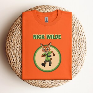 May include: Orange t-shirt featuring the character Nick Wilde from Zootopia. The design includes the text "NICK WILDE" in green above a cartoon image of Nick Wilde in a green circle. The shirt is folded on a woven, circular surface.