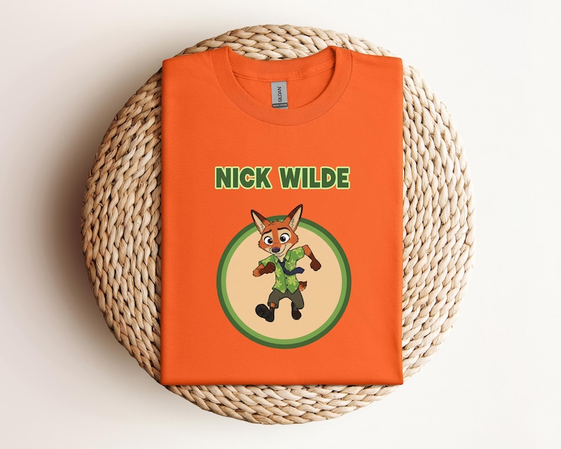 May include: Orange t-shirt featuring the character Nick Wilde from Zootopia. The design includes the text "NICK WILDE" in green above a cartoon image of Nick Wilde in a green circle. The shirt is folded on a woven, circular surface.