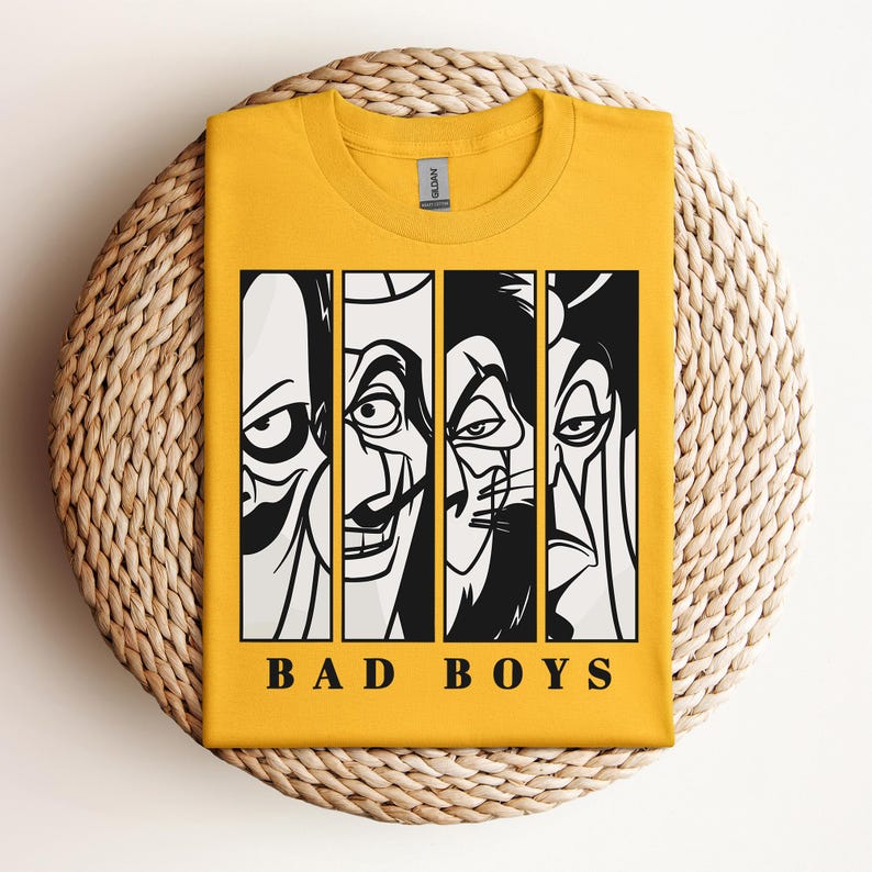 May include: Mustard yellow t-shirt featuring a graphic of four villainous faces in black and white, divided into vertical panels. Below the faces, the text "BAD BOYS" is printed in a bold, sans-serif font. The shirt is folded on a woven, circular surface.