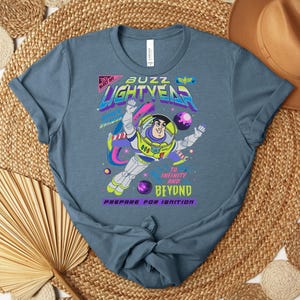 Puede incluir: Una camiseta azul con un dise&ntilde;o de Buzz Lightyear. El dise&ntilde;o incluye el texto "Buzz Lightyear" en una fuente retro colorida, junto con la frase "&iexcl;Hasta el infinito y m&aacute;s all&aacute;!".