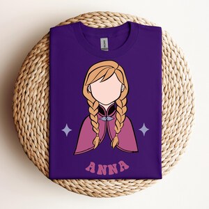 May include: A purple short-sleeved t-shirt featuring a graphic illustration of a character with blonde hair in two braids, wearing a pink cape with a silver clasp. The design includes two light purple four-pointed stars and the text ANNA in pink.