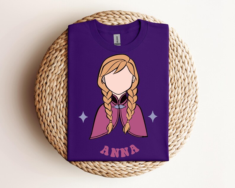 May include: A purple short-sleeved t-shirt featuring a graphic illustration of a character with blonde hair in two braids, wearing a pink cape with a silver clasp. The design includes two light purple four-pointed stars and the text ANNA in pink.