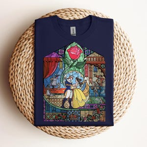 May include: Navy blue t-shirt featuring a stained-glass design with characters from a fairy tale. The design includes a red rose, a couple dancing, and other figures. The shirt is folded on a woven surface.