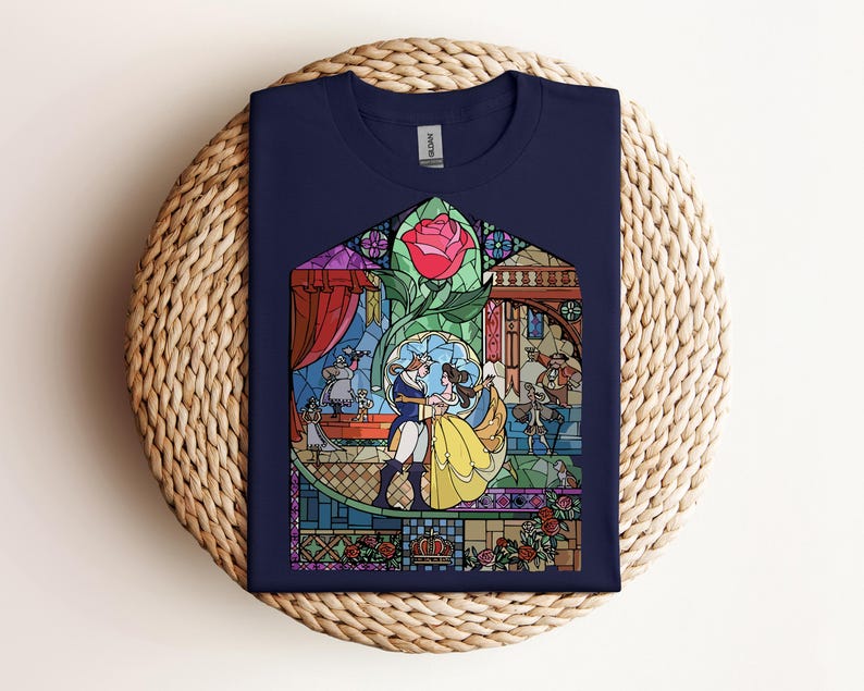 May include: Navy blue t-shirt featuring a stained-glass design with characters from a fairy tale. The design includes a red rose, a couple dancing, and other figures. The shirt is folded on a woven surface.