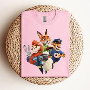 May include: A light pink t-shirt featuring a colorful graphic of characters from the animated movie Zootopia. The design includes a fox, a rabbit, a sheep, a cheetah, and other characters. The shirt is displayed on a woven, circular surface.