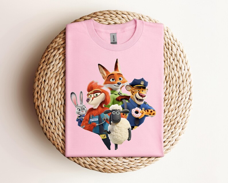 May include: A light pink t-shirt featuring a colorful graphic of characters from the animated movie Zootopia. The design includes a fox, a rabbit, a sheep, a cheetah, and other characters. The shirt is displayed on a woven, circular surface.