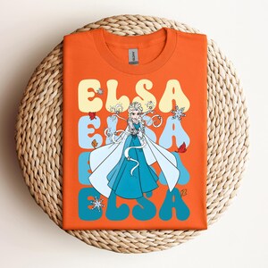 Op de afbeelding: Oranje t-shirt met het woord "ELSA" in een retro lettertype, met een illustratie van Elsa uit Frozen. Het ontwerp bevat sneeuwvlokken en herfstbladeren. Het shirt is op een geweven oppervlak gevouwen.